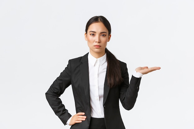 Serious-looking determined young asian businesswoman introduce her project, showing something on palm, holding hand right on blank space, as introduce product, standing in black suit white background