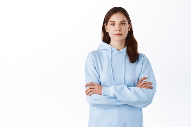 Serious-looking girl student in hoodie, cross arms on chest and look like professional with determined face, standing against white wall