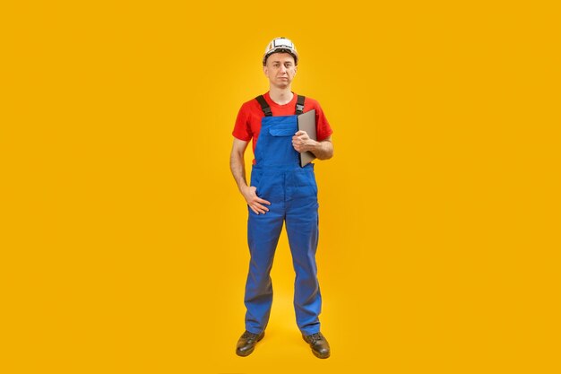 Serious male engineer in overalls and hardhat holding on clipboard and looking at camera isolated on yellow background Copy space