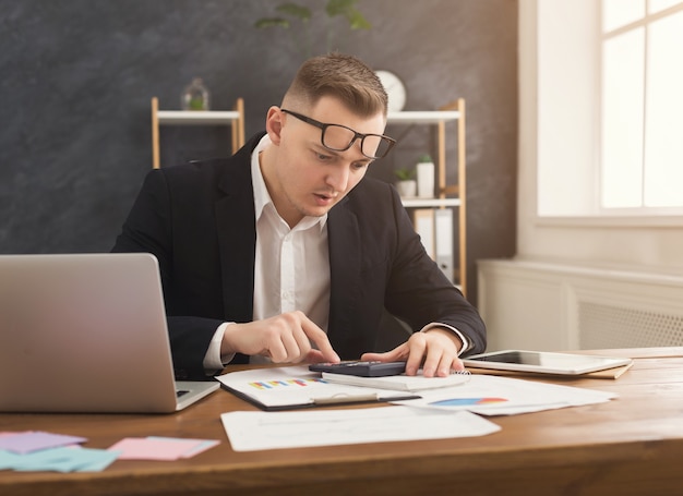 Photo serious male financial advisor working with papers and laptop at modern office interior. pensive man in formal wear checking documents and thinking over strategy, copy space