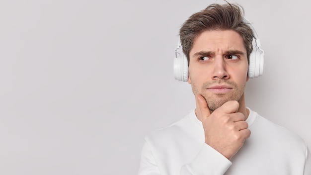 Serious male student listens educational content holds chin wears wireless headphones with good sound quality on ears looks away with attentive expression isolated over white wall copy space