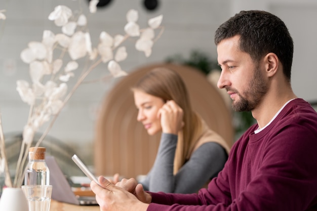 Serious man in casualwear scrolling in touchpad while sitting in cafe with young woman