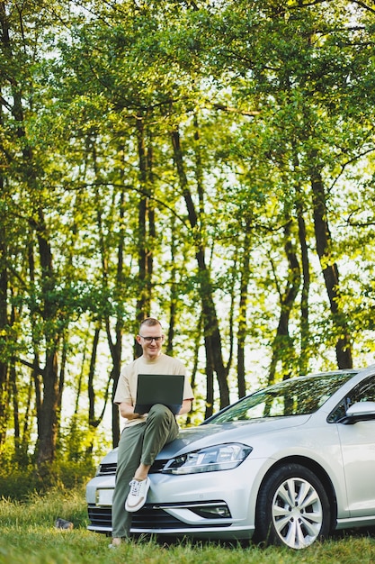 A serious man in glasses is working on a laptop on the hood of his car Remote work in nature Work on a laptop online Working on a computer while traveling