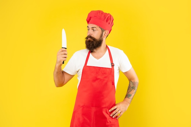 Serious man in red apron and toque looking at chefs knife yellow background, chef