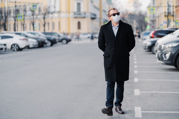 serious man walks at street, dressed in elegant outfit, wears medical mask to prevent coronavirus or another type of virus, stays safe during quarantine time. Pandemic situation