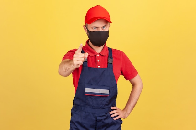 Serious man in worker uniform and protective mask showing finger looking at camera