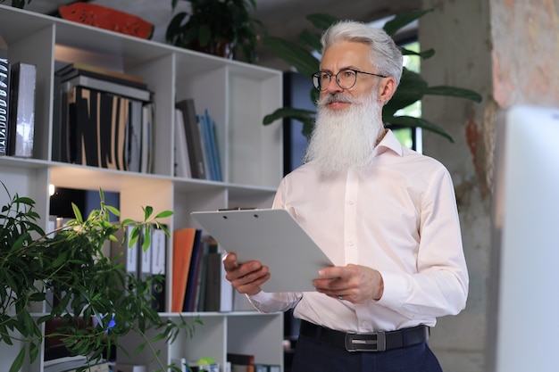 Serious mature business man in white shirt reading contract while standing in the office.