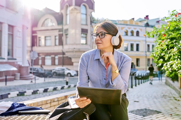 Serious mature business woman in glasses headphones with digital tablet on city street