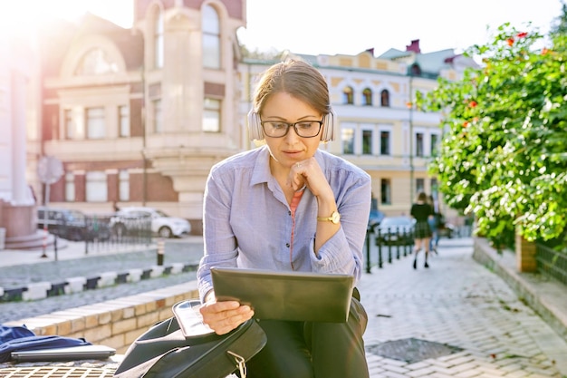 Serious mature business woman in glasses headphones with digital tablet on city street