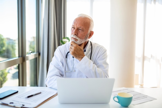 Serious mature doctor using laptop and sitting at desk Senior professional medic physician wearing white coat and stethoscope working on computer at workplace