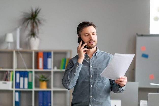 Serious middle aged businessman working at office standing by workdesk holding documents and having