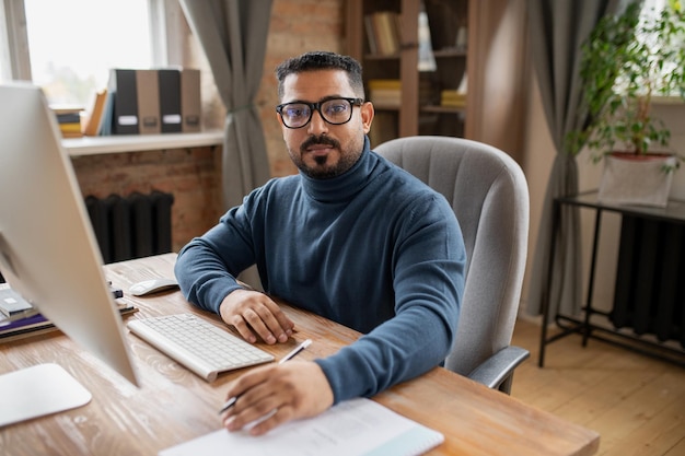 Serious middle aged teacher looking at camera while sitting by desk in front of computer monitor in home environment