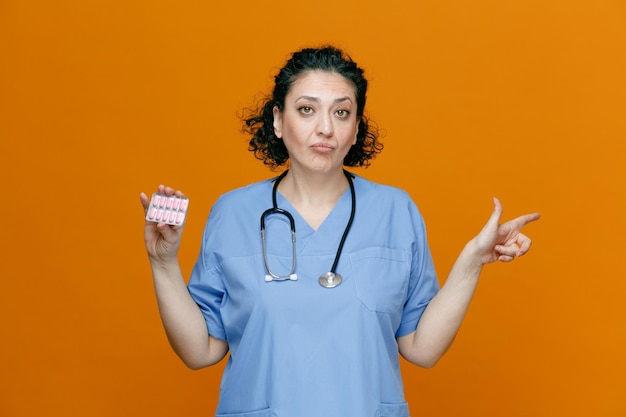 Serious middleaged female doctor wearing uniform and stethoscope around her neck looking at camera pointing to side showing pack of capsules isolated on orange background