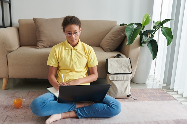 Serious mixed race girl in glasses and ear buds sitting with crossed legs on floor and making notes in workbook while learning lesson online