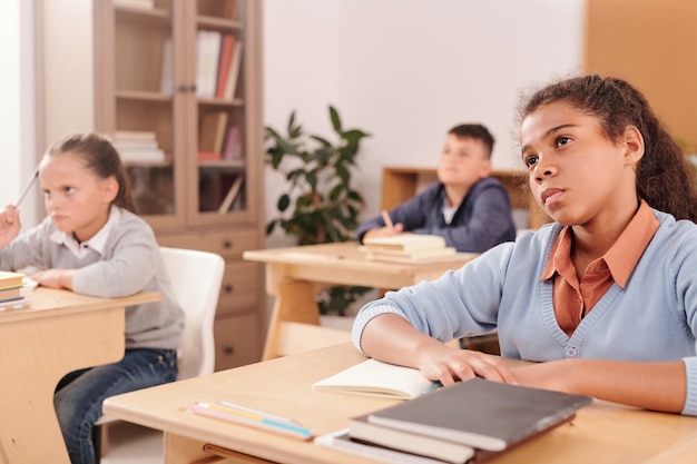 Serious mixed-race student of secondary school and her classmates looking at teacher attentively and listening to her explanation at lesson