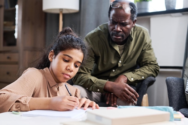 Serious multiethnic schoolgirl making notes in copybook while doing homework