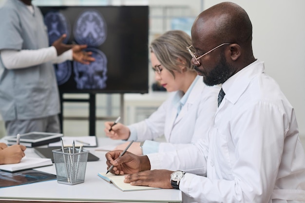 Serious professional doctors sitting at table and making notes during medical conference in office