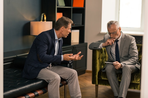 Serious psychotherapist sitting in his office while listening to his worried client and looking at notes in his notepad