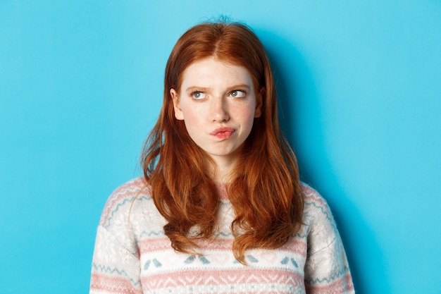 Serious redhead girl thinking, grimacing and frowning while pondering, looking left at copy space, standing over blue background.