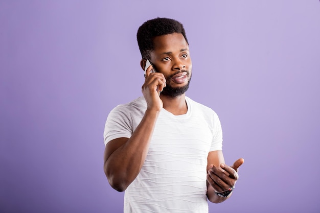 Serious sad african american man talking on mobile phone, wearing white tshirt looking up with thoughtful dissatisfied expression on violet background. Face expressions and communication concept.