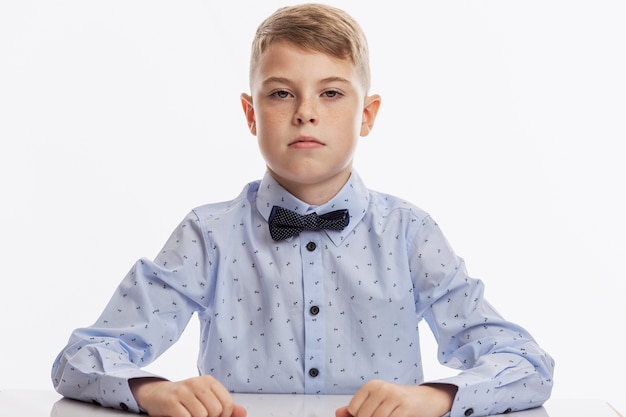 serious schoolboy in a blue shirt with a bow tie sits at the table.