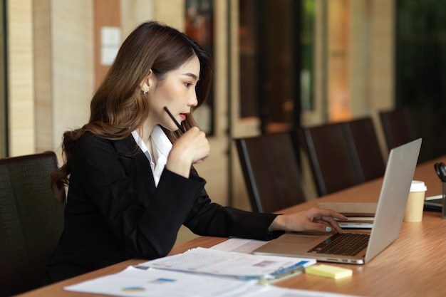 Serious smart young businesswoman touching face with pen working with her laptop business report