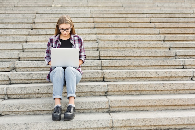 Serious student girl sitting on stairs working with laptop, preparing for exams outdoors, having rest in university campus. Technology, education and remote working concept, copy space