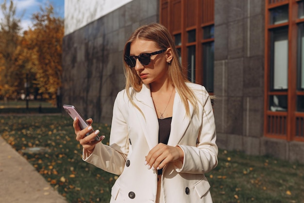 Serious stylish businesswoman in sunglasses and white suit browsing internet on smartphone in city