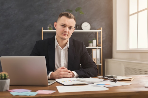 Serious successful male financial advisor working with papers and laptop at modern office. Pensive man in formal wear checking documents and thinking over strategy, looking at camera, copy space