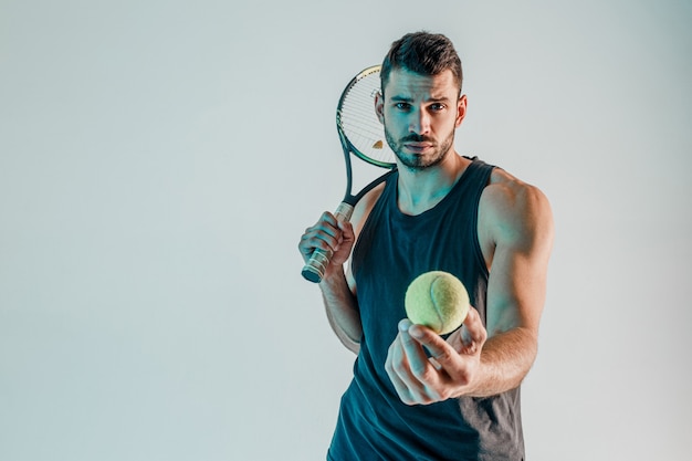 Serious tennis player hold racquet and showing ball at camera. Front view of young bearded european sportsman looking at camera. Isolated on turquoise background. Studio shoot. Copy space