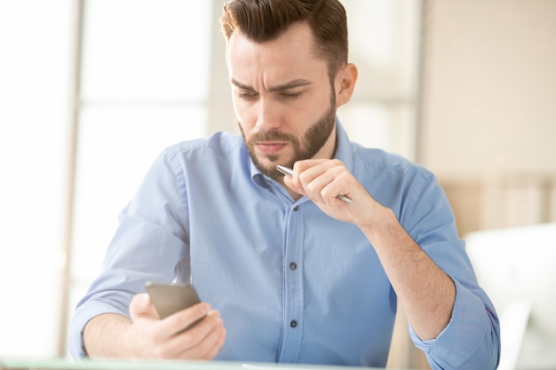 Serious or tense young office manager reading message or promo in smartphone that he cannot understand