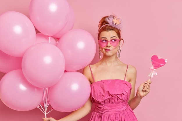 Serious thoughtful redhead woma purses lips thinks about celebration wears trendy sunglasses and dress holds sweet candy and balloons prepares for graduation party