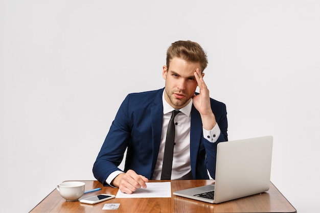 Serious and tired young workaholic, businessman in office feeling dizzy and distressed, working all day, sitting with laptop, documents and coffee, touching temple, have headache