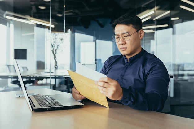 Serious and upset asian businessman reading letter with bad news investor man working inside modern