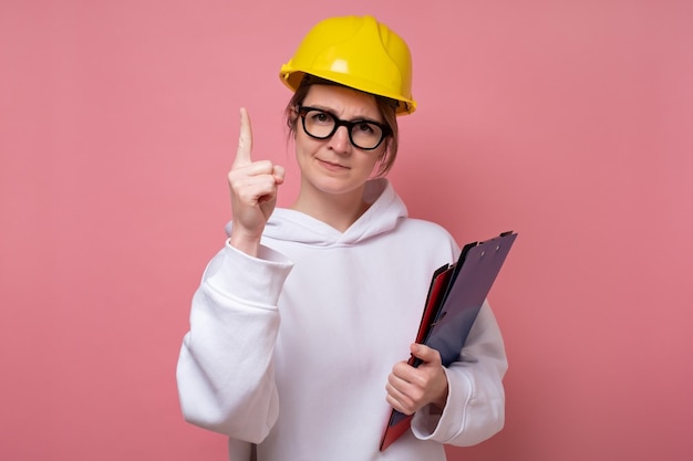 Serious woman in the yellow helmet points up giving advice. Studio shot on pink wall.