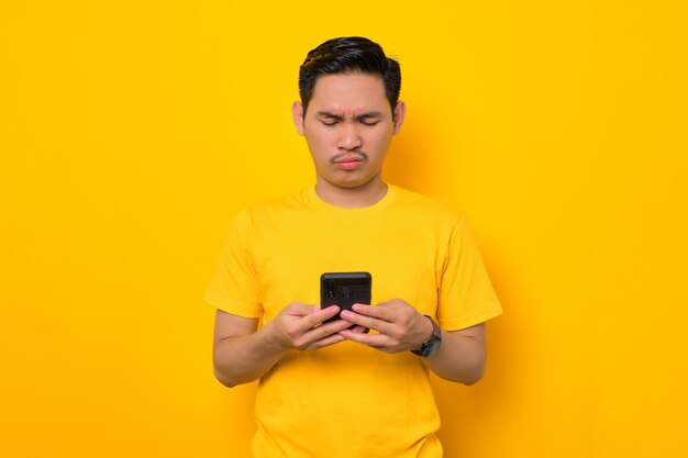 Serious young Asian man in casual tshirt using mobile phone and looking at gadget screen isolated on yellow background People lifestyle concept