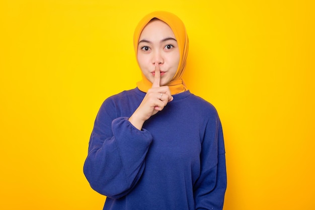 Serious young Asian Muslim woman dressed in casual sweater showing shh gesture asks to be quiet isolated over yellow background