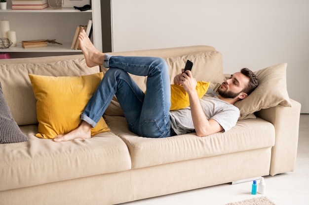 Serious young bearded man in jeans lying on sofa and communicating online via smartphone messengers in home isolation