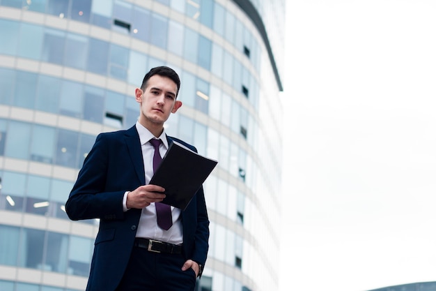 Serious young businessman in suit holding documents, folder in hands and looking at camera outdoors