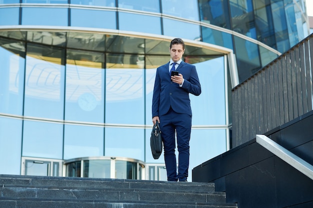 Serious young businessman with case tapping message outside business center