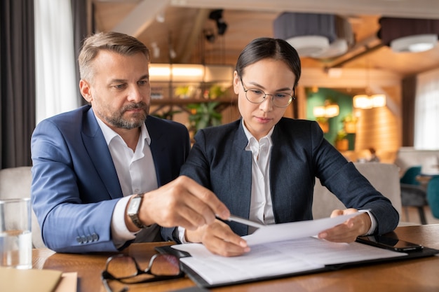 Serious young businesswoman looking through paper before signing while her employer explaining its main points