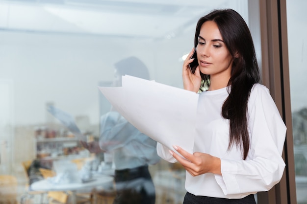 Serious young businesswoman working with documents and talking on mobile phone in office