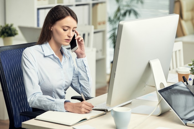 Serious young caucasian businesswoman making notes in planner and talking by phone while working in ...