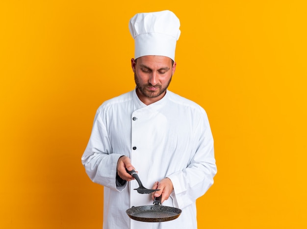 Serious young caucasian male cook in chef uniform and cap holding frying pan and spatula above it looking at them 