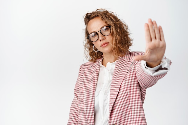 Serious young corporate woman stretch out hand in stop sign, showing prohibition, permission denied. Businesswoman forbid something bad, white background.