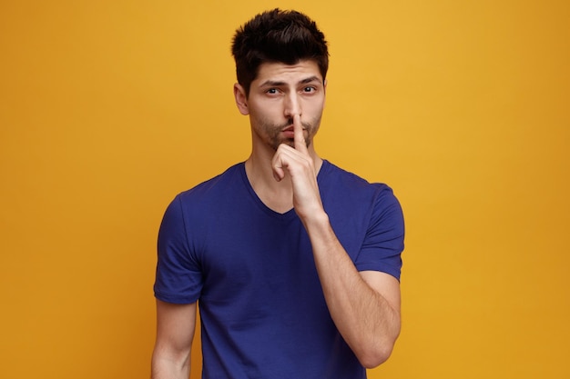 Serious young handsome man looking at camera doing silence gesture on yellow background