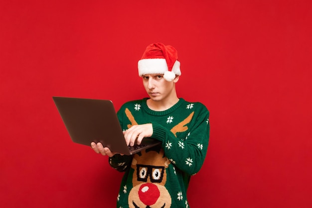 Serious young man in christmas sweater and hat santa stands with laptop on red background