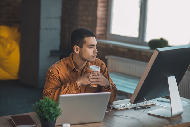 Serious young man looking at the laptop screen while sitting with a cup of coffee