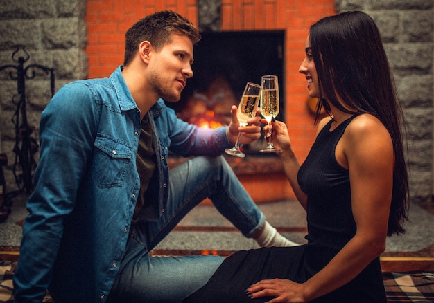 Serious young man sit on floor with his girlfriend and look at her. She smiles to him. They hold glasses with champagne. Couple sit at fireplace.