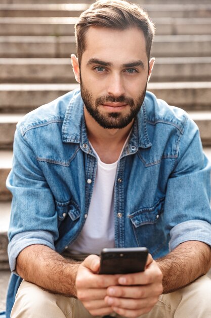 Serious young man sitting on stairs outdoors, using mobile phone